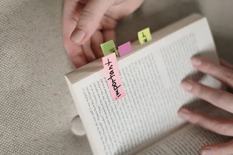 Tangan memegang buku terbuka dengan nota pelekat merah jambu dan hijau.