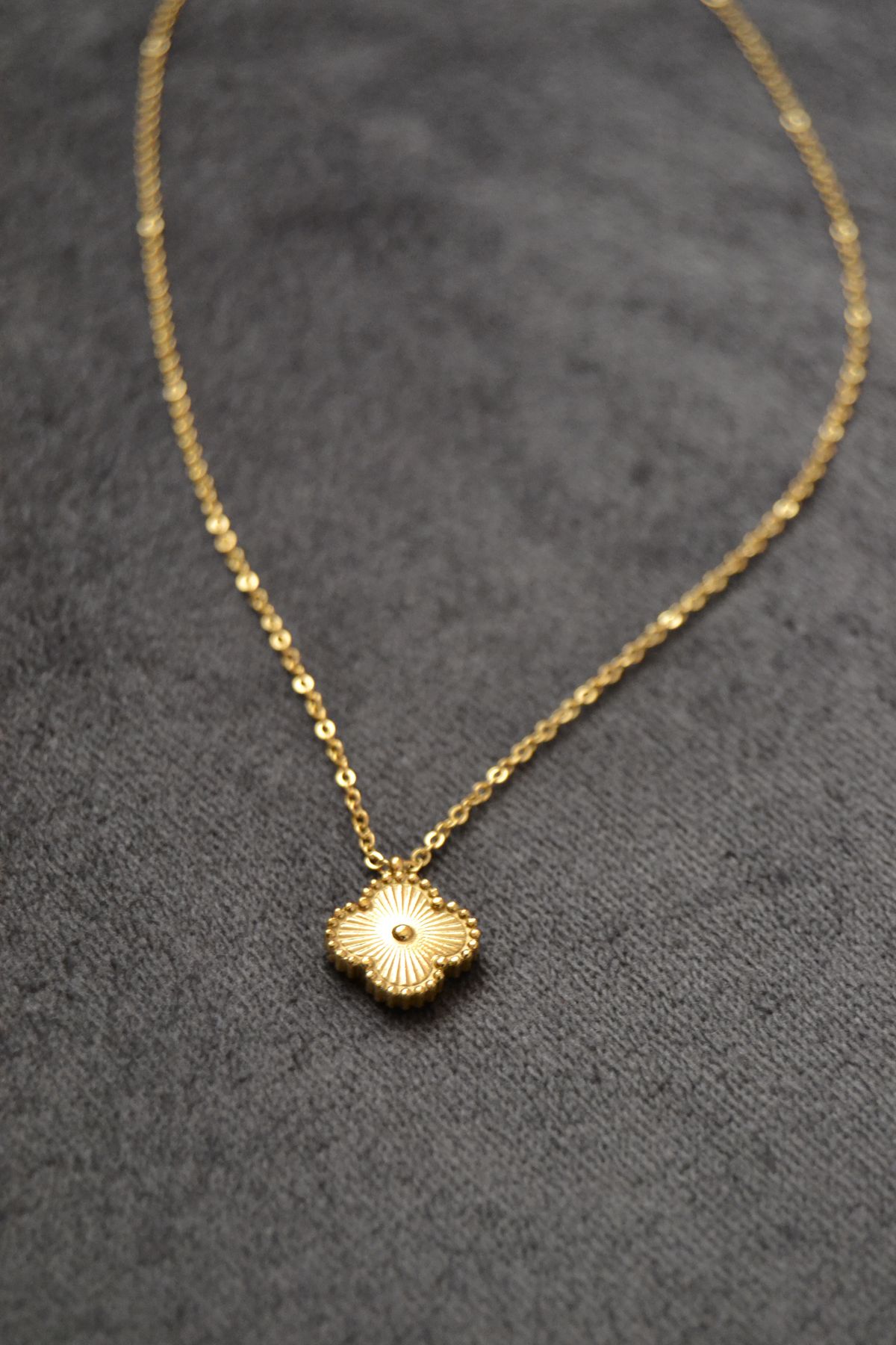 A gold-coloured necklace with a flower-shaped pendant rests on grey fabric.